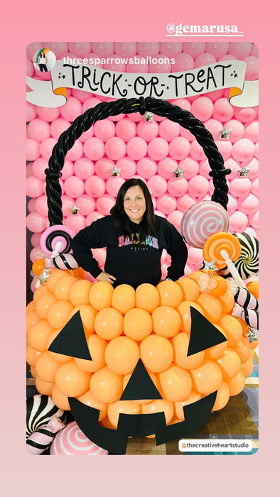 Jenny Porter from Three Sparrows poses inside a large balloon pumpkin bucket photo op, featuring an oversized jack-o-lantern made of balloons with black face details, balloon candy decorations, and a pink balloon wall backdrop with a “Trick or Treat” banner. A festive Halloween balloon display perfect for parties, schools, and event installations.
