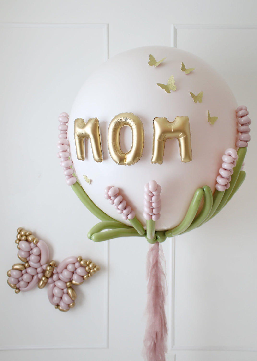 Elegant jumbo Mother's Day balloon featuring gold "MOM" letters, delicate pink balloon flowers, green stems, and butterfly accents. Perfect for gifting or party décor, this design adds a stunning touch to Mother’s Day celebrations.