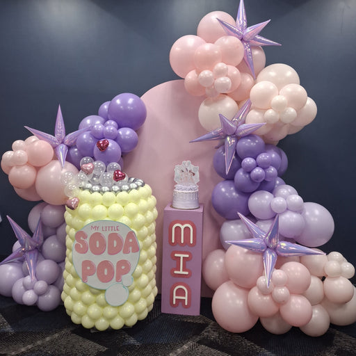 K-pop Demon Hunters inspired balloon setup featuring a pastel soda pop can made of balloons with a “My Little Soda Pop” label, a pink bottle prop with the name “MIA,” and clusters of pink, purple, and cream balloons accented with metallic lavender stars, perfect for K-pop Demon Hunters themed parties and pop culture celebrations.