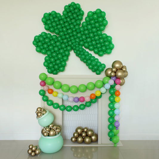 Large green linking balloon shamrock displayed above a white fireplace with layered rainbow balloon garland and gold chrome balloon clusters. Includes a mint green pot of gold balloon prop with metallic gold balloons spilling out, styled by Lilly Jimenez for St Patrick’s Day party decor, balloon tutorials, and festive event backdrops.