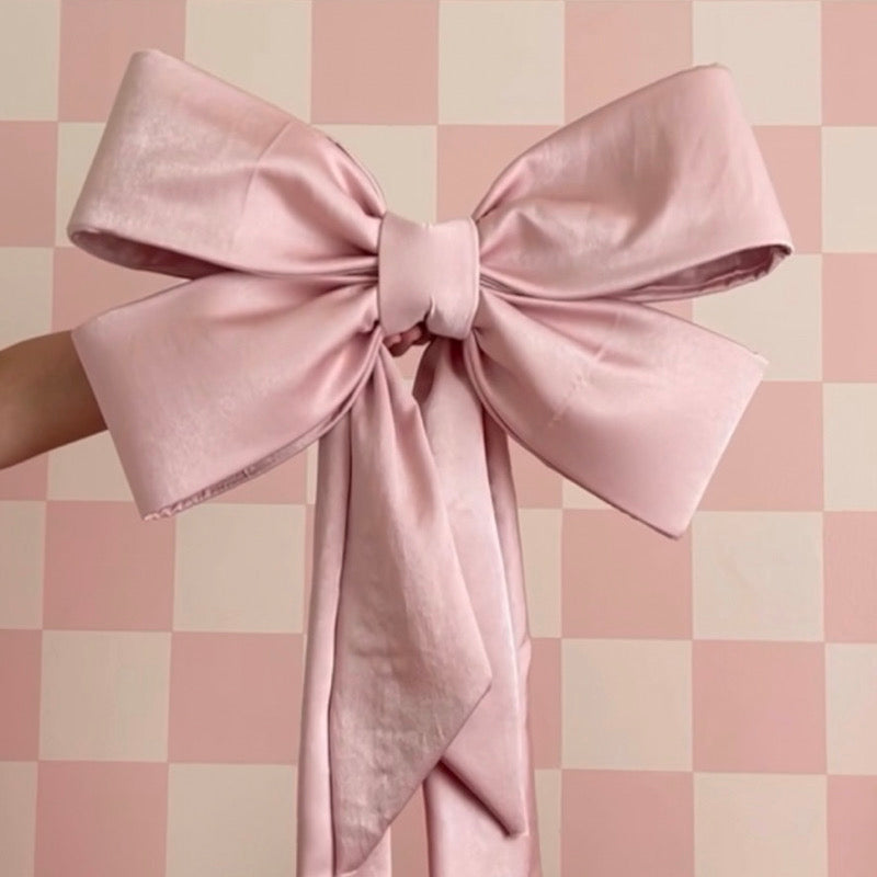 A beautifully crafted oversized pink fabric bow with long trailing ribbons, held against a pink and cream checkered background. The bow features smooth, structured loops and a neatly wrapped center, showcasing an elegant and polished design.
