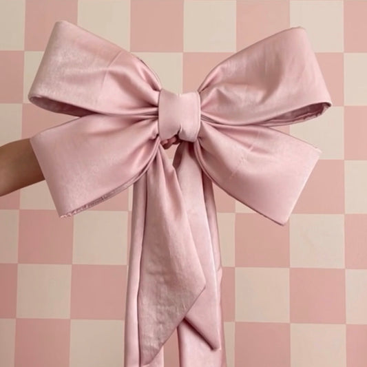 A beautifully crafted oversized pink fabric bow with long trailing ribbons, held against a pink and cream checkered background. The bow features smooth, structured loops and a neatly wrapped center, showcasing an elegant and polished design.