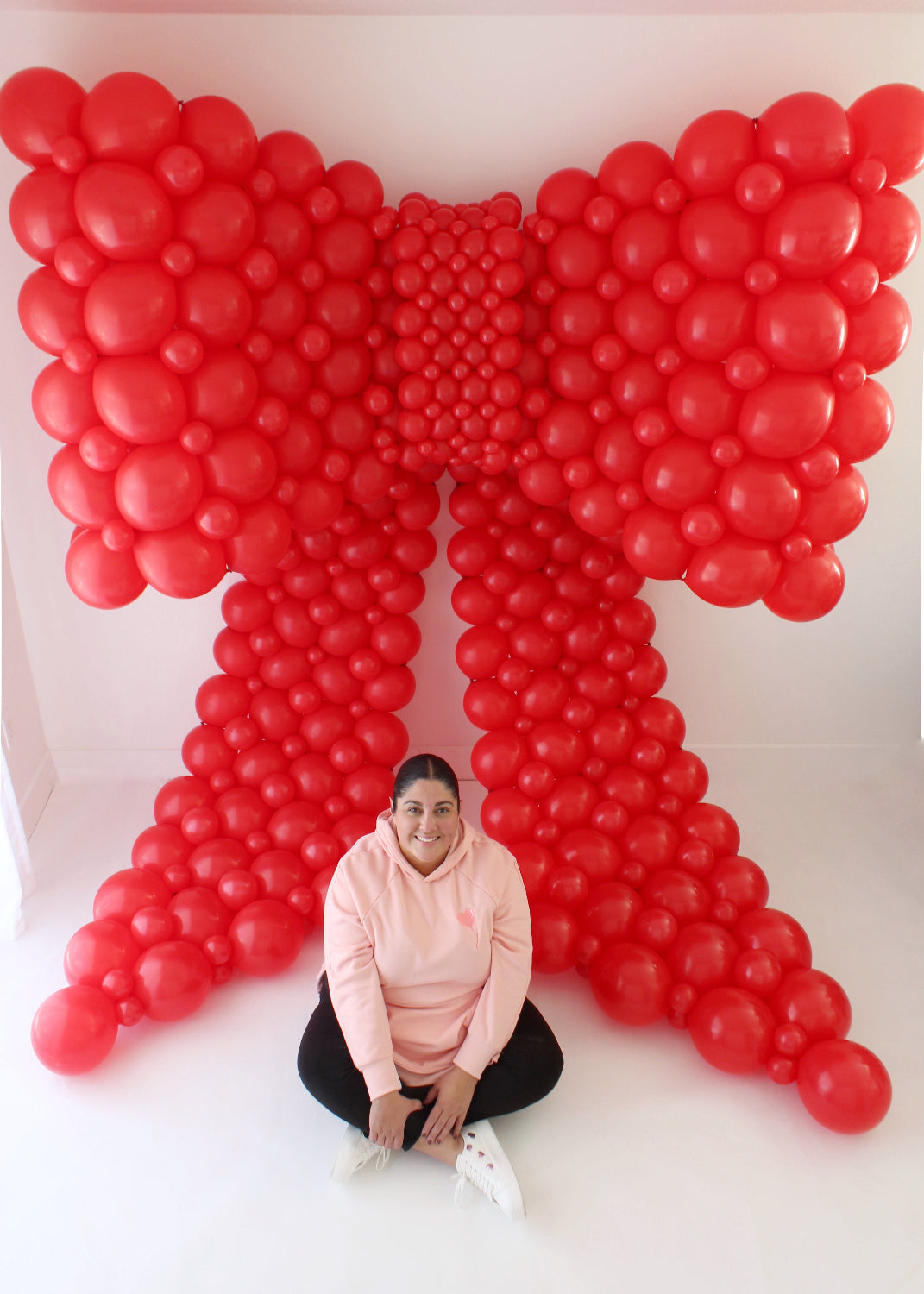 Lilly Jimenez sitting in front of an oversized red linking balloon bow, showcasing the scale and structure of the bow's loops and tails. The rounded bubble technique highlights what students learn in the Linking Balloon Bows Class.