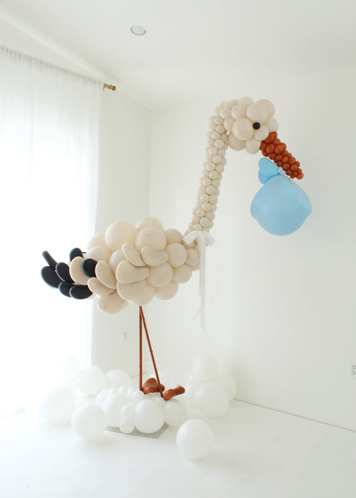 Large stork balloon display created with soft neutral balloons, featuring a tall curved neck, cloud style base, and a blue balloon bundle held in the beak, designed as a welcome baby statement piece for baby showers, newborn arrivals, nursery decor, and styled photography setups.