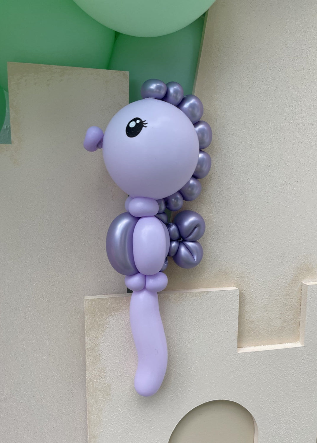Lavender balloon seahorse with metallic purple details and a sculpted tail, featured in the Under the Sea Balloon Class by Lilly Jimenez. Displayed against a foam cutout backdrop, this balloon sculpture is perfect for under-the-sea, mermaid, or ocean-themed parties. The class includes video instruction on how to recreate this seahorse using twisting and layering techniques.