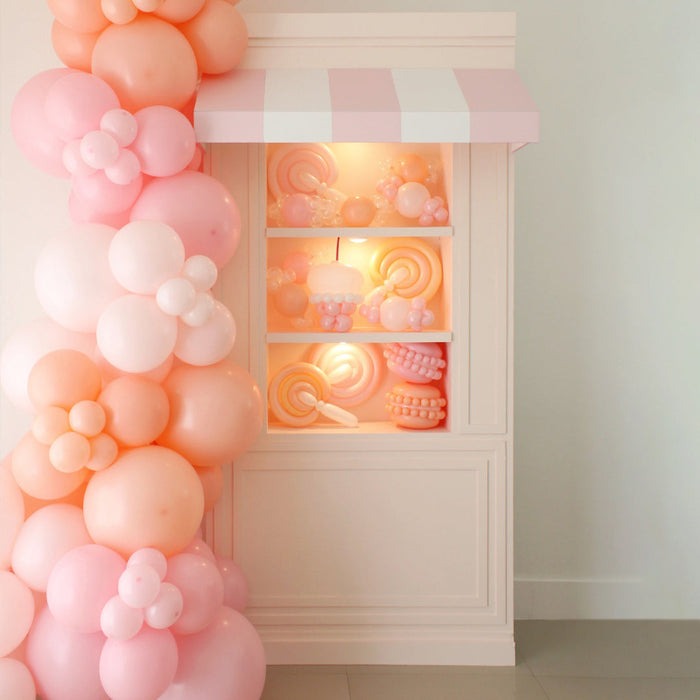 Pink and white balloon arch in front of a decorative window with candy-themed lights.