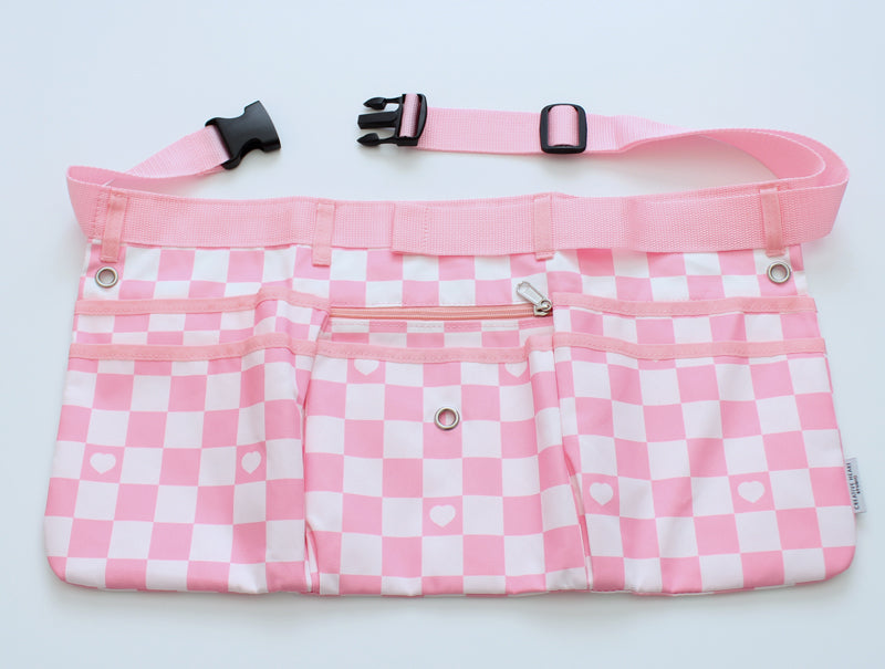 Light pink and white checkered maker apron with heart detail, featuring six pockets, a center zip pouch, belt loops, and grommets for attaching tools. Includes an adjustable strap with a side-release buckle for a secure and comfortable fit. Designed for balloon artists, event decorators, and creative professionals, this stylish and functional workbelt offers ample storage for tools and accessories.
