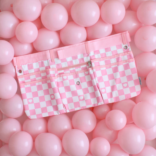 Light pink and white checkered maker apron with heart detail, displayed on a vibrant background of pink balloons. Features multiple pockets, a center zip pouch, belt loops, and grommets for tool attachments. Designed for balloon artists, event decorators, and creative professionals, this stylish and functional workbelt offers ample storage space and an adjustable fit for comfort during events and installations.