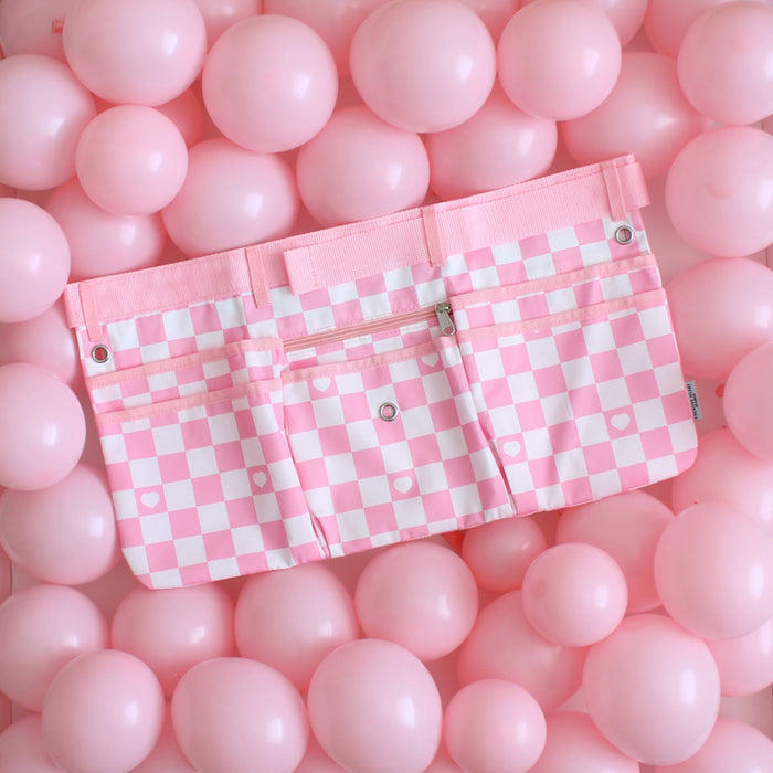 Light pink and white checkered maker apron with heart detail, displayed on a vibrant background of pink balloons. Features multiple pockets, a center zip pouch, belt loops, and grommets for tool attachments. Designed for balloon artists, event decorators, and creative professionals, this stylish and functional workbelt offers ample storage space and an adjustable fit for comfort during events and installations.