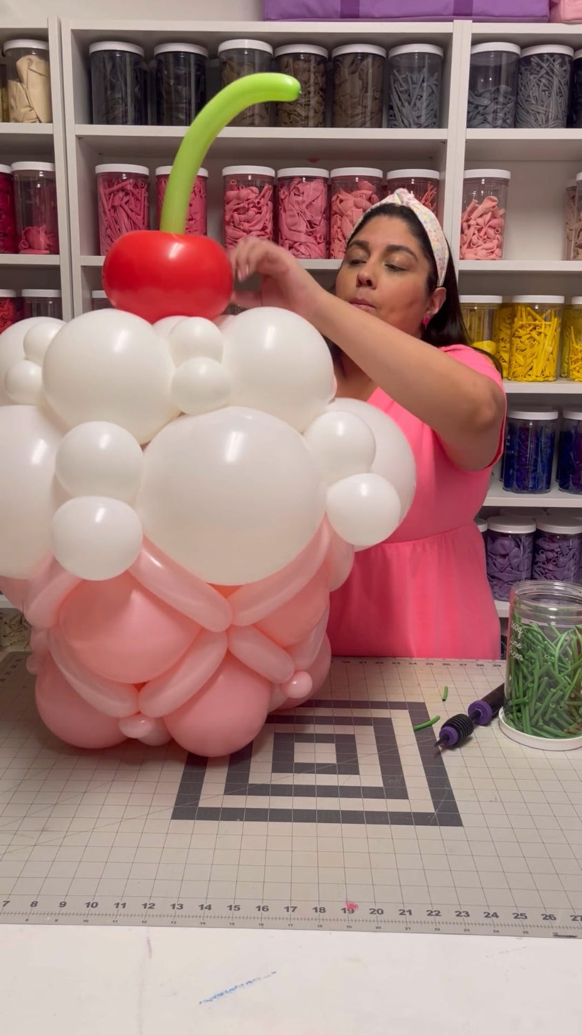 Lilly works on assembling a large balloon cupcake with pink frosting layers and a white whipped topping, topped with a red balloon cherry and green balloon stem. The balloon sculpture is part of her Linking Balloon Cupcake Tutorial, ideal for birthdays, candyland themes, dessert bars, and eye-catching party displays.