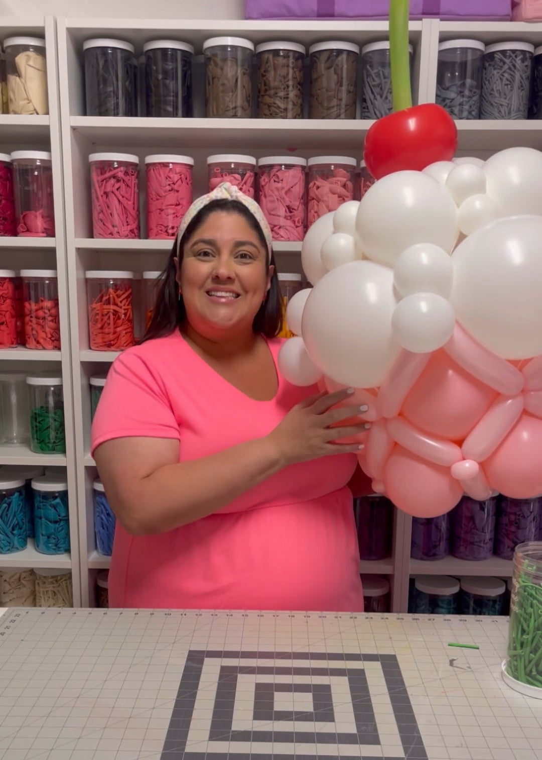 Lilly smiles while holding an oversized balloon cupcake featuring pink and white frosting details, topped with a red balloon cherry and green stem. This playful and whimsical design is part of her Linking Balloon Cupcake Tutorial, perfect for birthdays, candy-themed parties, and balloon decorators looking for fun statement pieces.