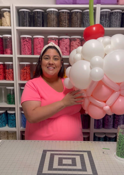 Lilly smiles while holding an oversized balloon cupcake featuring pink and white frosting details, topped with a red balloon cherry and green stem. This playful and whimsical design is part of her Linking Balloon Cupcake Tutorial, perfect for birthdays, candy-themed parties, and balloon decorators looking for fun statement pieces.