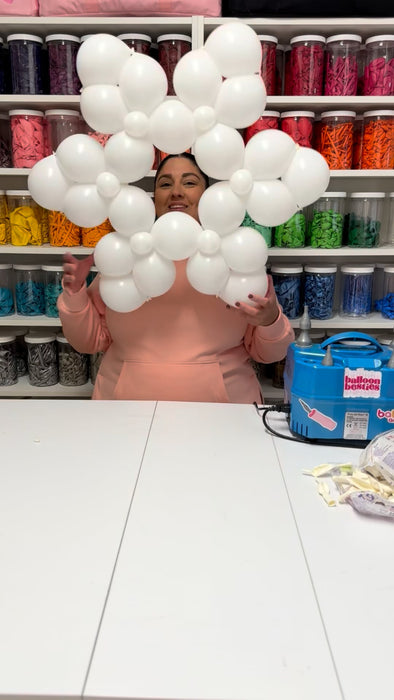 Lilly stands in her studio wearing a light pink hoodie, holding a large white balloon snowflake made from clusters of small round balloons. Behind her is a wall of clear jars filled with balloons sorted by color, creating a bright organized backdrop. A blue electric balloon pump sits on the table beside scattered balloon scraps, giving a behind the scenes look at her winter balloon-decor class.