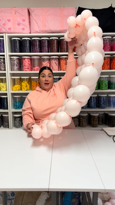 Lilly holding a curved blush balloon tail section while demonstrating how to shape and assemble the components of a linking balloon bow. The rounded cluster technique is clearly visible, reflecting skills taught in the class.