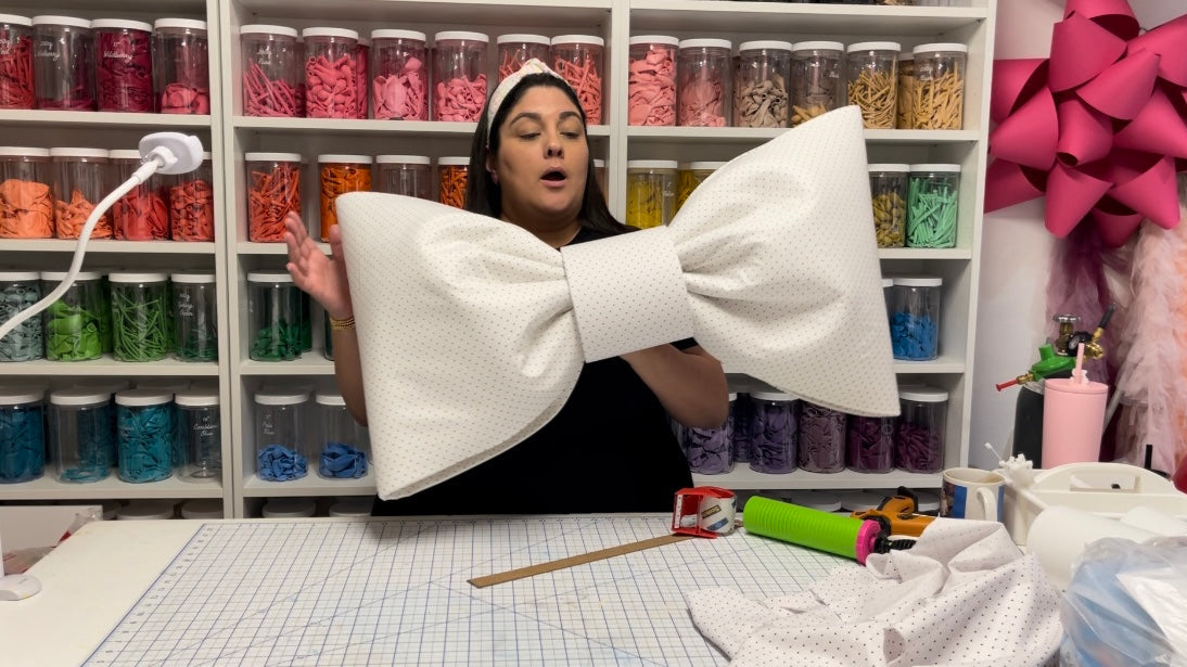 Lilly Jimenez stands in her craft studio, holding a large white polka-dotted bow while demonstrating a technique. Behind her, shelves filled with colorful balloons in clear jars line the walls. A cutting mat, ruler, tape, and crafting tools are spread across the workspace, showcasing the creative process of making oversized bows.