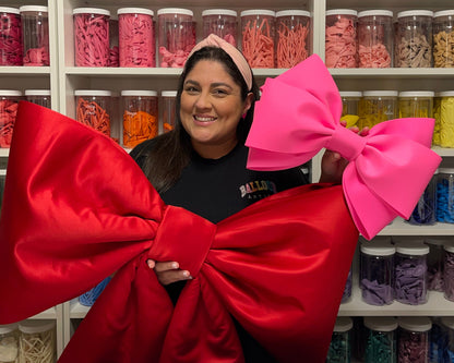 Lilly Jimenez stands in front of a wall of organized balloon supplies, smiling while holding two oversized bows—a large red satin bow and a smaller bright pink bow. The background features neatly arranged jars filled with colorful balloons, showcasing the variety of materials used in her creative designs.