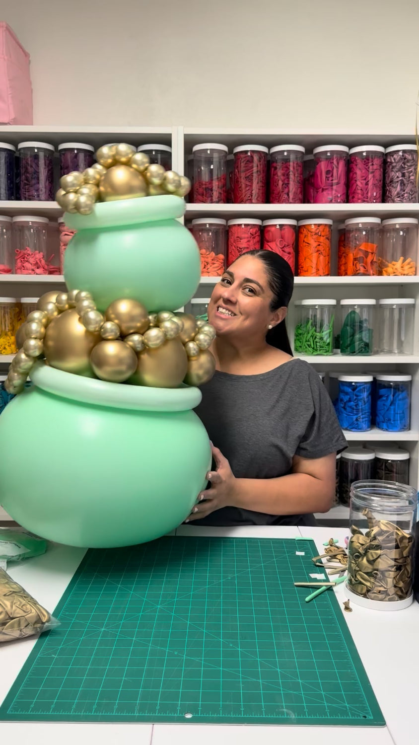 Lilly Jimenez holding a large mint green pot of gold balloon prop filled with metallic gold chrome balloons in her studio. Shelves of colorful balloon supplies line the background, showcasing a St Patrick’s Day balloon tutorial setup for party decor, balloon artists, and DIY event styling inspiration.