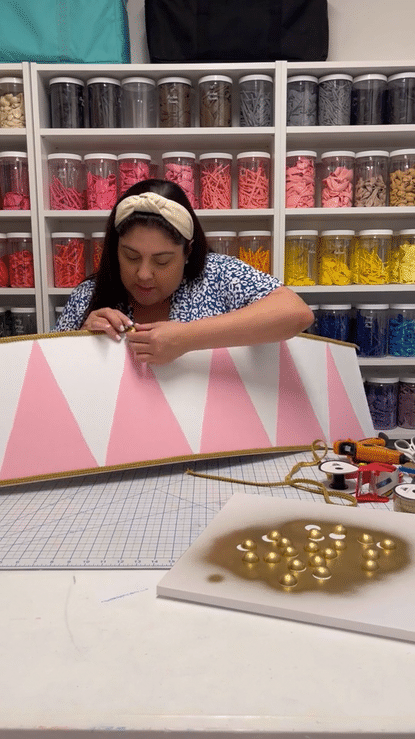 Lilly Jimenez carefully adds gold accents to a handmade Christmas drum panel in this DIY holiday balloon decor tutorial. The foam board panel features pink triangles and gold rope detailing, designed to wrap around balloon Christmas trees. Surrounding her are crafting tools, ribbon, and painted embellishments, with organized balloon jars in rainbow colors behind her. This behind-the-scenes moment captures the holiday magic and attention to detail Lilly brings to each Creative Heart Studio project.