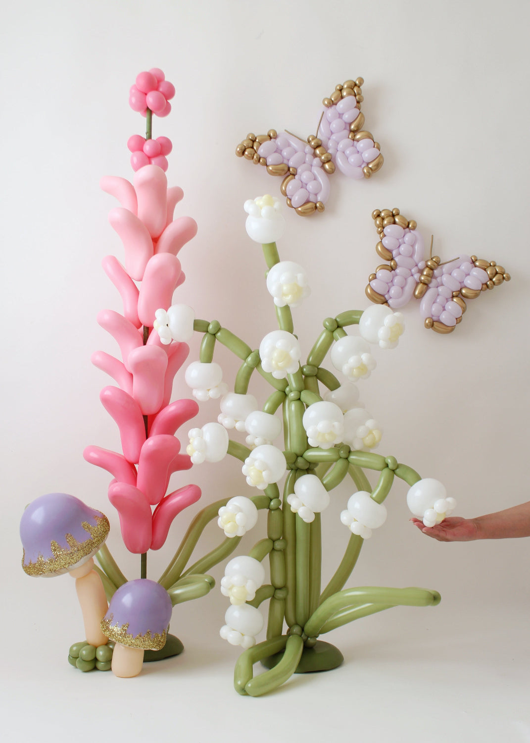 A stunning balloon display by Lilly Jimenez featuring lilies of the valley, pink lavender, and gold-detailed butterflies. The arrangement includes purple glitter-topped balloon mushrooms, adding a whimsical touch. Lilly’s hand is seen making final adjustments, highlighting her expertise in balloon artistry. Perfect for enchanted garden themes, weddings, and event decor inspiration.
