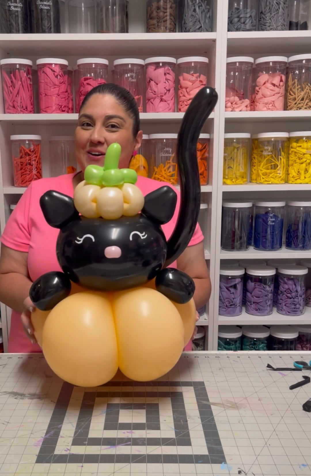 Lilly Jimenez of The Creative Heart Studio showcases an adorable black balloon cat sitting on a pumpkin-shaped balloon base, complete with a tiny pumpkin hat. Behind her, shelves filled with colorful balloons highlight her creative workspace. This fun and festive design is part of her Balloon Pumpkin & Cat Tutorial, perfect for Halloween, fall festivals, and seasonal decor.