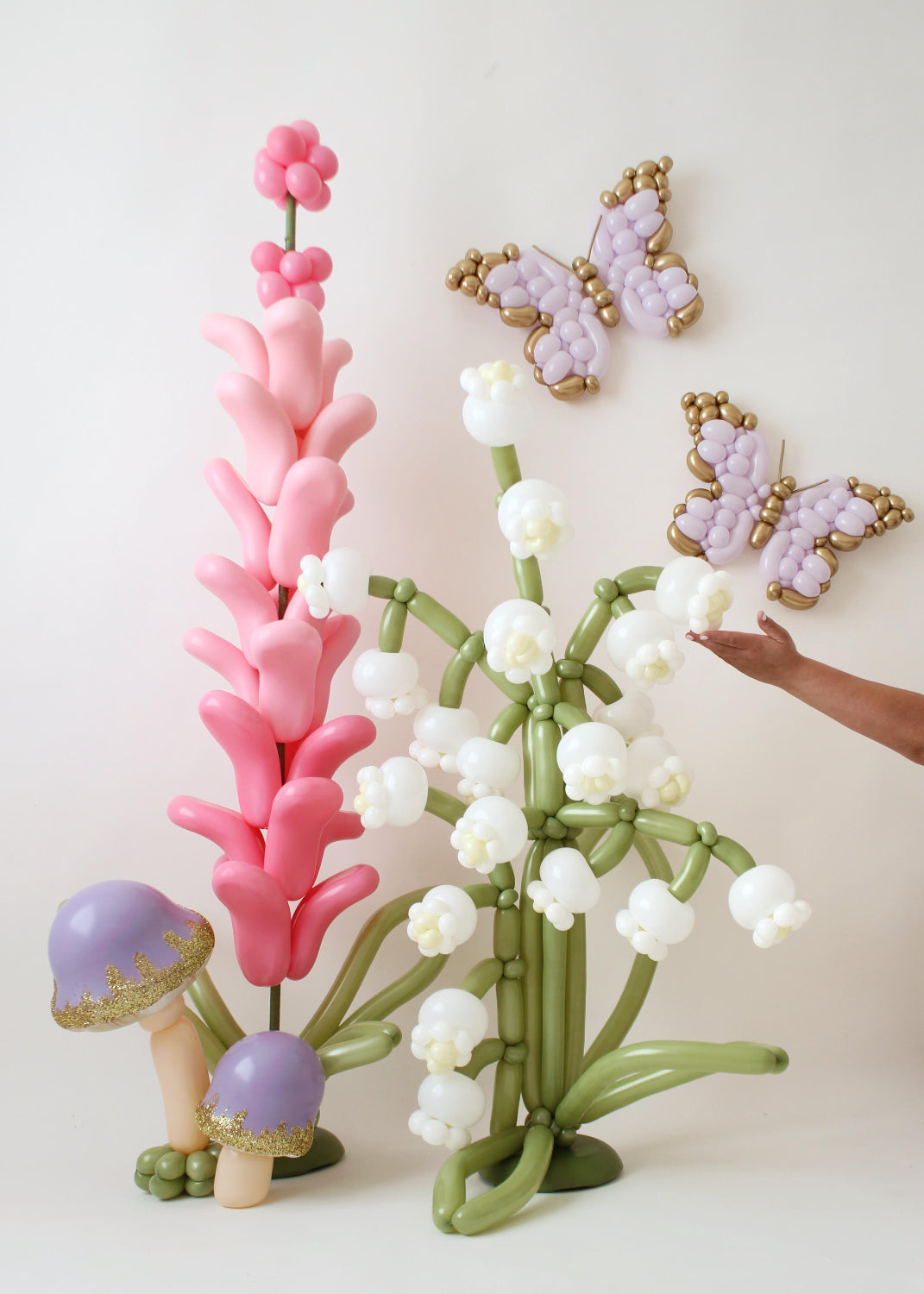 A whimsical balloon arrangement by Lilly Jimenez featuring oversized lilies of the valley, pink lavender, and golden-accented butterflies. The display includes glitter-topped balloon mushrooms, creating a magical, fairy-tale-inspired scene. Lilly's hand is seen adjusting the delicate details, showcasing her artistry in balloon decor. Perfect for enchanted garden themes, event styling, and balloon art inspiration.