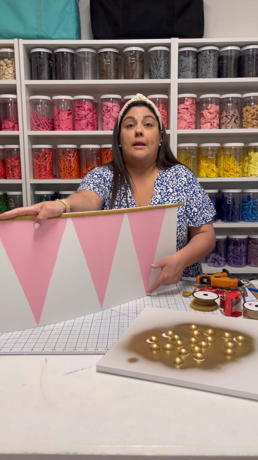 Lilly Jimenez from Creative Heart Studio demonstrates how to make a festive Christmas drum base using foam board in her holiday balloon tutorial. She holds a pink and white drum panel trimmed with gold rope, perfect for wrapping around balloon trees. The background features a vibrant rainbow wall of balloon jars, while the table shows materials like ribbon, glue, and painted gold accents. A behind-the-scenes look at crafting holiday decor with professional, polished results.