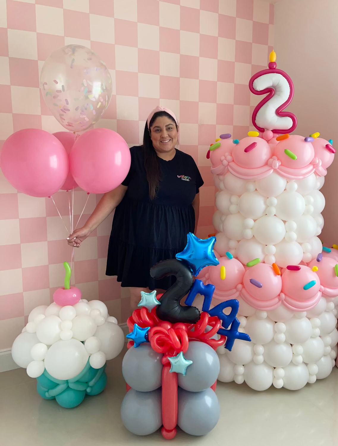 Lilly Jimenez stands in front of a pink checkered backdrop, surrounded by creative balloon birthday decorations. To her right is a towering two-tiered balloon cake with pink frosting details, colorful sprinkles, and a number "2" topper. To her left, she holds a cluster of pink and confetti-filled balloons next to a cupcake-shaped balloon sculpture. In the foreground, a red, blue, and silver balloon arrangement displays the numbers "2," "1," and "4," adding a festive touch.