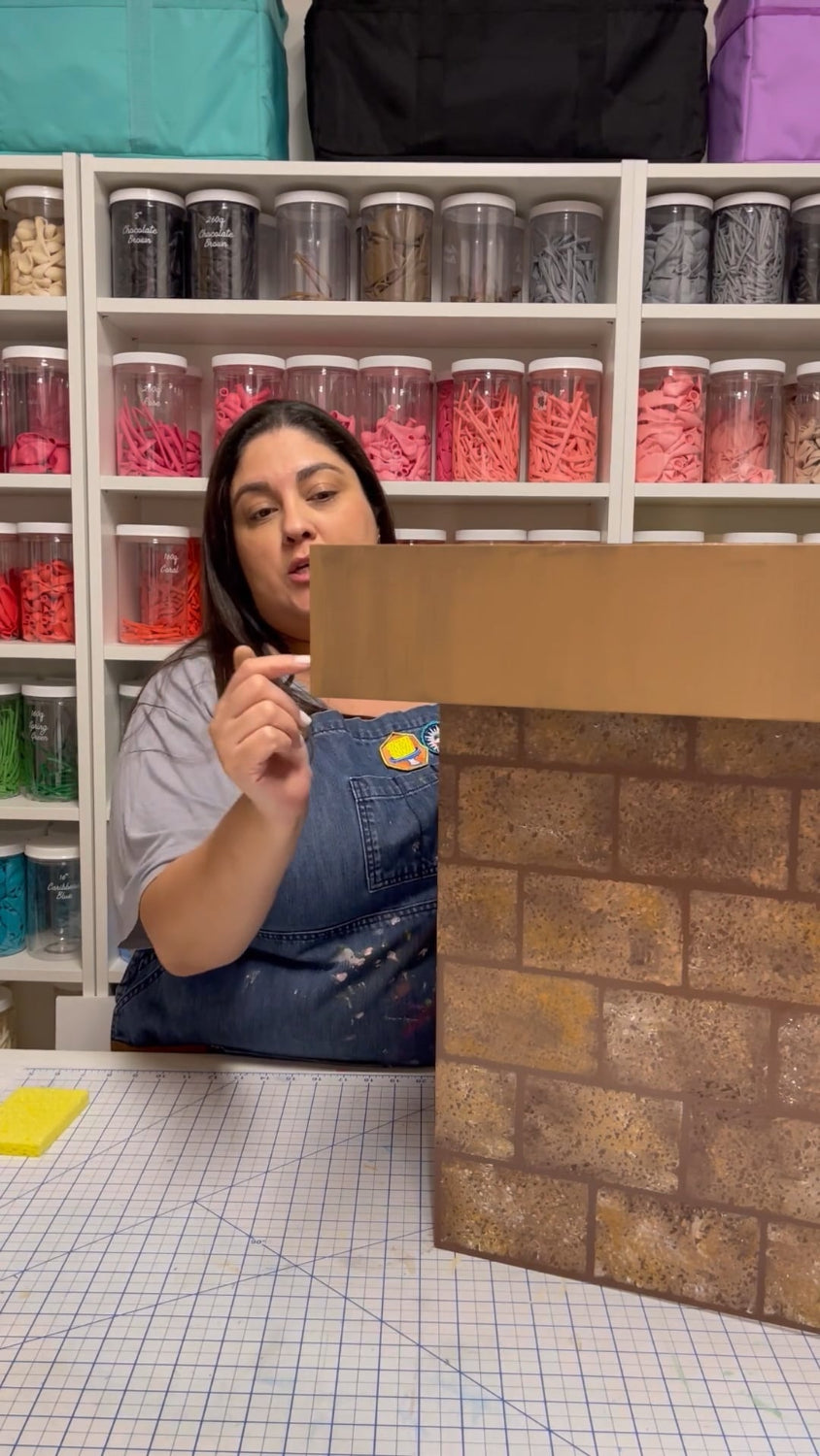 Lilly Jimenez demonstrates how to paint a realistic brick pattern on foamboard for her DIY Santa Chimney tutorial. She stands in her colorful balloon-filled studio, teaching step-by-step holiday decor techniques perfect for Christmas photo setups and party props.