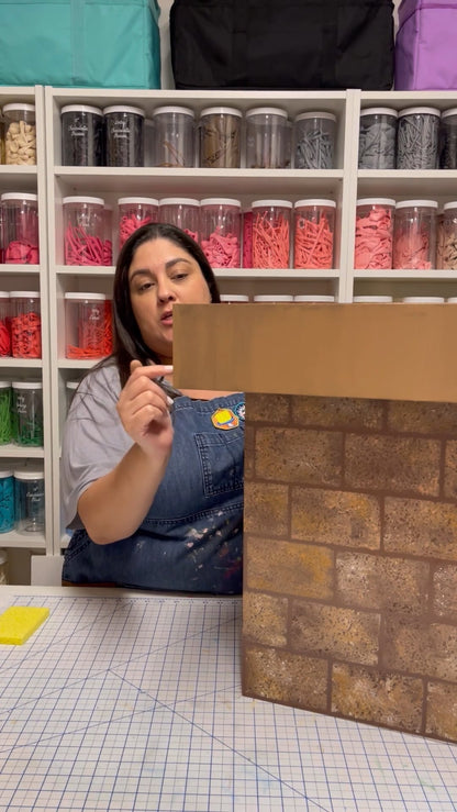Lilly Jimenez demonstrates how to paint a realistic brick pattern on foamboard for her DIY Santa Chimney tutorial. She stands in her colorful balloon-filled studio, teaching step-by-step holiday decor techniques perfect for Christmas photo setups and party props.