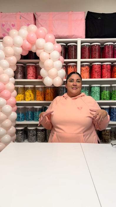 Lilly stands in her studio wearing a light pink hoodie, demonstrating how to build a pastel pink and white balloon candy cane column. Behind her is a wall of organized balloon jars arranged by color, along with pink storage bags on the top shelf. A partially built candy cane balloon structure curves into the frame on the left, creating a bright and creative workspace scene perfect for balloon-decor tutorials.