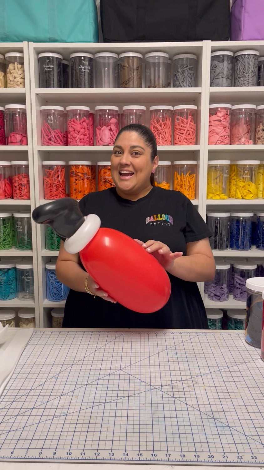 Lilly Jimenez smiling while holding a red and black balloon leg prop from her Santa Chimney tutorial. Behind her is a rainbow-organized wall of balloons, showcasing her festive and fun workspace for holiday DIY classes.