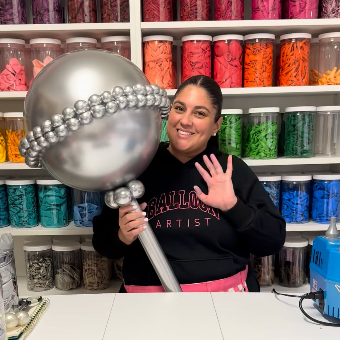 Lilly Jimenez holding an oversized silver balloon baby rattle prop inside her balloon studio, surrounded by organized jars of balloons. This balloon structure highlights a professional prop-building technique used for baby showers, gender reveals, and themed balloon decor, as taught in Creative Heart Studio balloon classes.