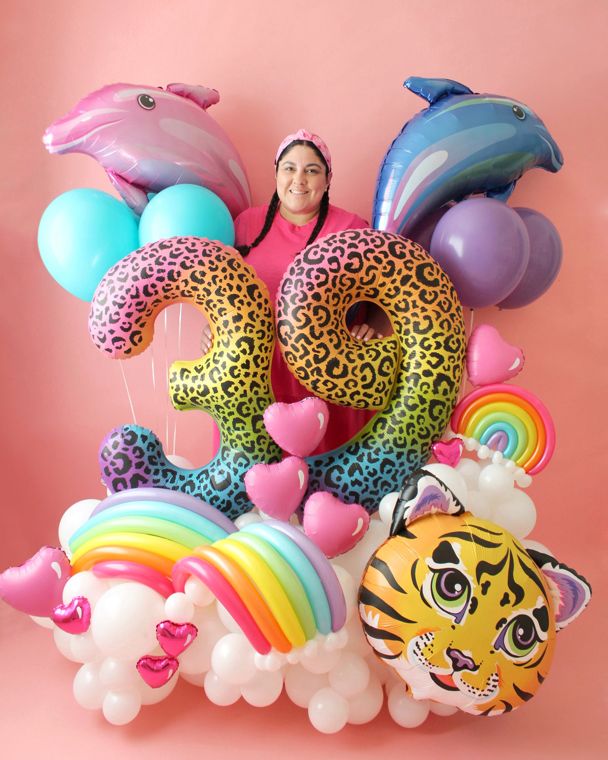 Lilly Jimenez poses with her vibrant Lisa Frank-inspired birthday balloon arrangement. The display features large spray-painted foil number balloons "39" in a colorful leopard print, a tiger face balloon, rainbow balloon arches, pink heart balloons, and two dolphin foil balloons. Set against a soft pink background, this whimsical and nostalgic balloon bouquet embodies the playful and bold aesthetic of Lisa Frank.