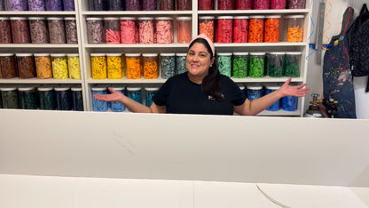 Lilly Jimenez stands behind a large foam board panel while preparing to build the Sand Castle Backdrop. Surrounded by shelves filled with color-coded balloon jars, this workspace highlights the creative setup for her DIY balloon and foam board class, available at The Creative Heart Studio.