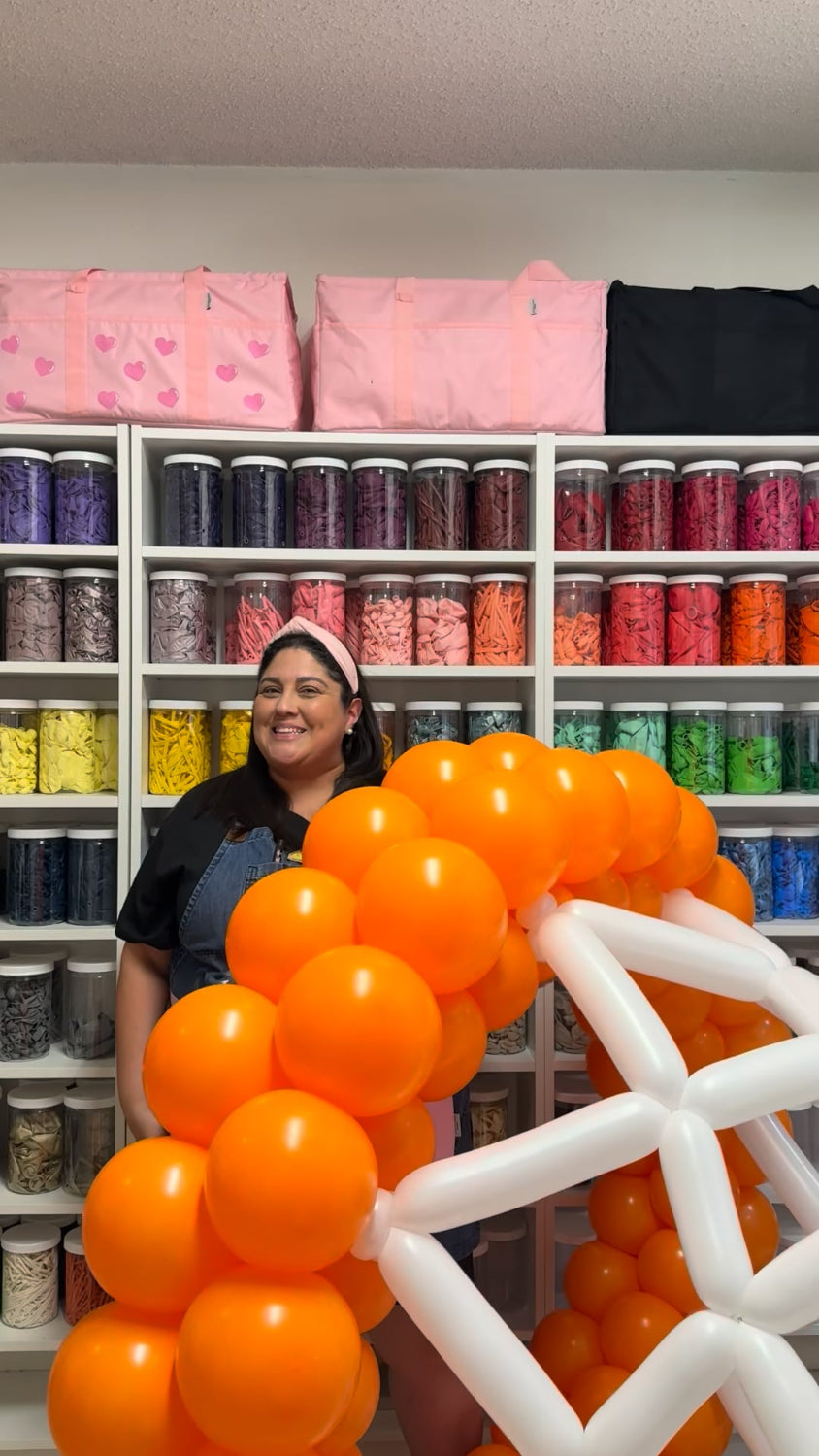 Lilly Jimenez poses with an oversized balloon basketball hoop in her studio, surrounded by a rainbow wall of balloon supplies. The hoop features bright orange and white balloons, part of the Oversized Basketball Hoop Balloon Sculpture Class. Perfect behind-the-scenes look at the creative process from The Creative Heart Studio.