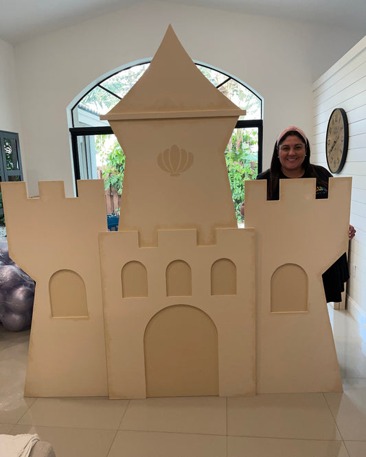 Lilly Jimenez showcases the completed full-size foam board Sand Castle Backdrop in a bright indoor space. The castle structure includes arched windows, a turret with a shell design, and decorative battlements. This project is featured in her on-demand class from The Creative Heart Studio and works for beach, princess, or fairytale-themed parties.