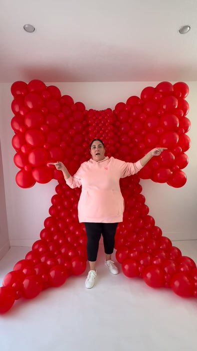 Lilly standing in front of a large red linking balloon bow with arms outstretched, demonstrating the full height and width of the bow and the rounded bubble-linking technique taught in the class.