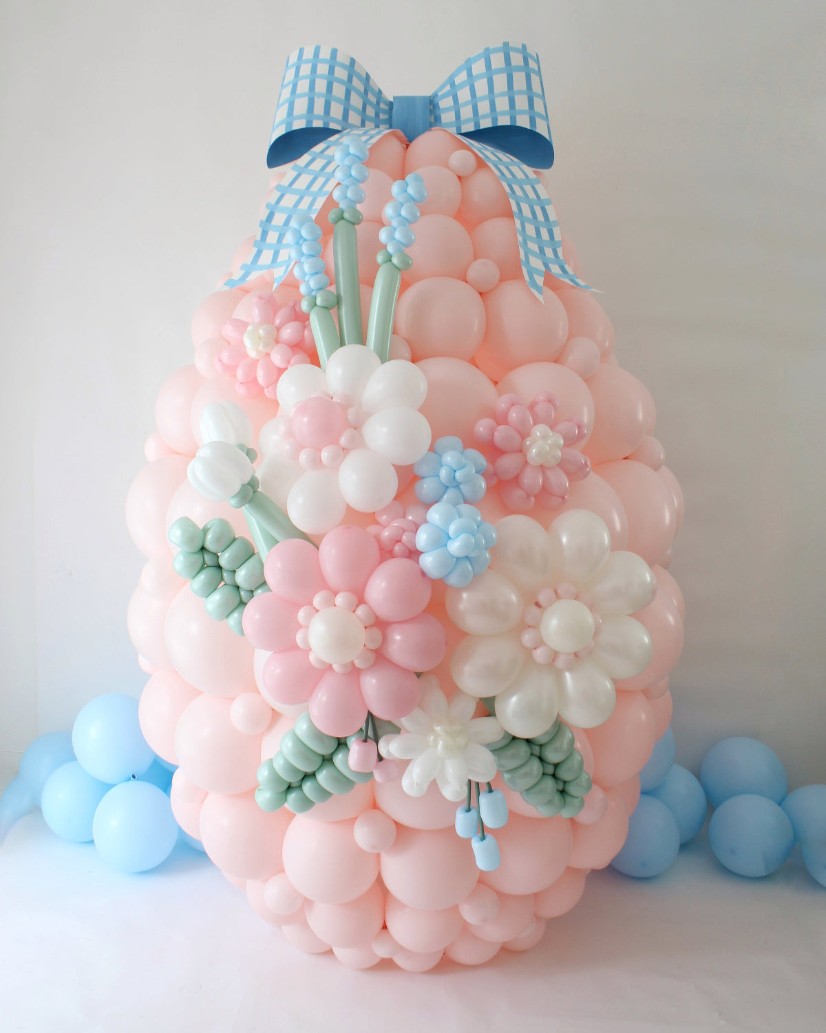 Large pastel pink balloon Easter egg made with linking balloons and decorated with balloon flowers, greenery, and a blue gingham bow. A stunning spring centerpiece created in Lilly’s Easter Balloon Class.