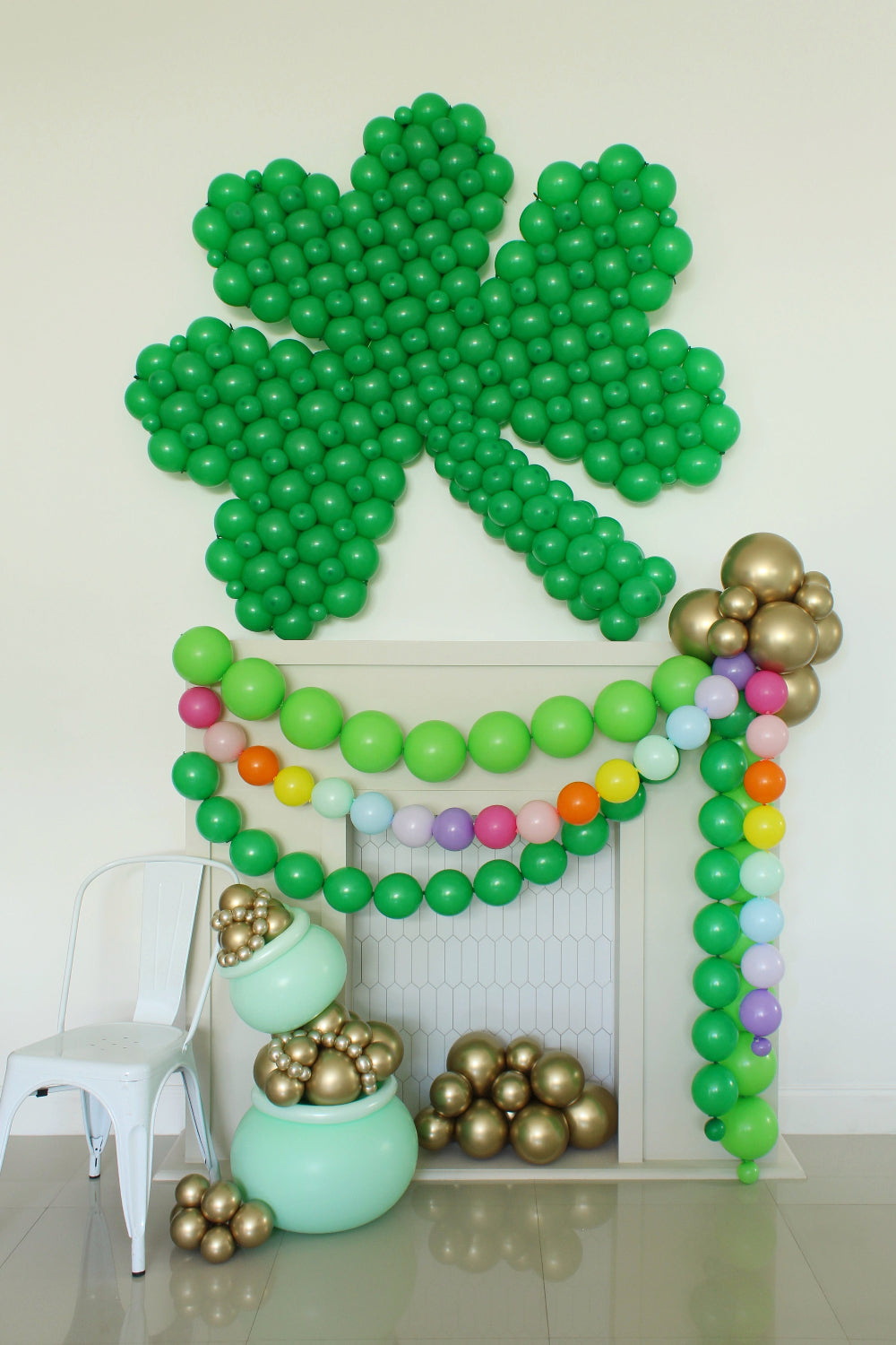 Large linking balloon shamrock design mounted above a white fireplace backdrop with a pot of gold balloon arrangement and rainbow garland, created by Lilly Jimenez for St Patrick’s Day balloon decor. Features rich green balloons forming a clover, gold chrome balloon clusters, and pastel rainbow accents for festive party styling and photo backdrops.