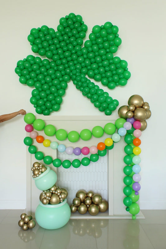 Oversized linking balloon shamrock in rich green mounted above a white fireplace backdrop with rainbow balloon garland and gold chrome balloon clusters. Features pastel rainbow accents, a mint green pot of gold balloon arrangement, and festive St Patrick’s Day balloon decor styled by Lilly Jimenez for party backdrops and event installations.