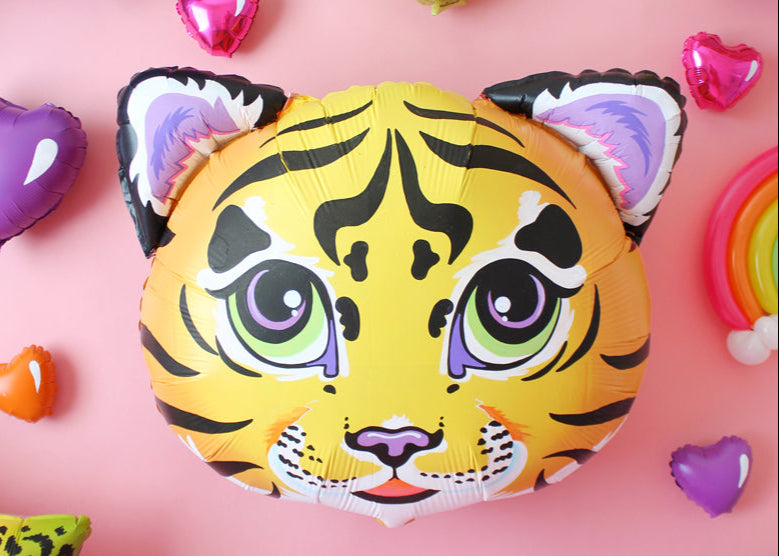 A customized, hand-painted tiger face foil balloon with bright green and purple eyes, bold black stripes, and pastel ear details, inspired by the iconic Lisa Frank aesthetic. Set against a soft pink background, this unique balloon is surrounded by vibrant heart-shaped foils in pink, purple, orange, and blue, along with a partial rainbow balloon. A nostalgic, colorful creation that’s every ‘90s girl’s dream!