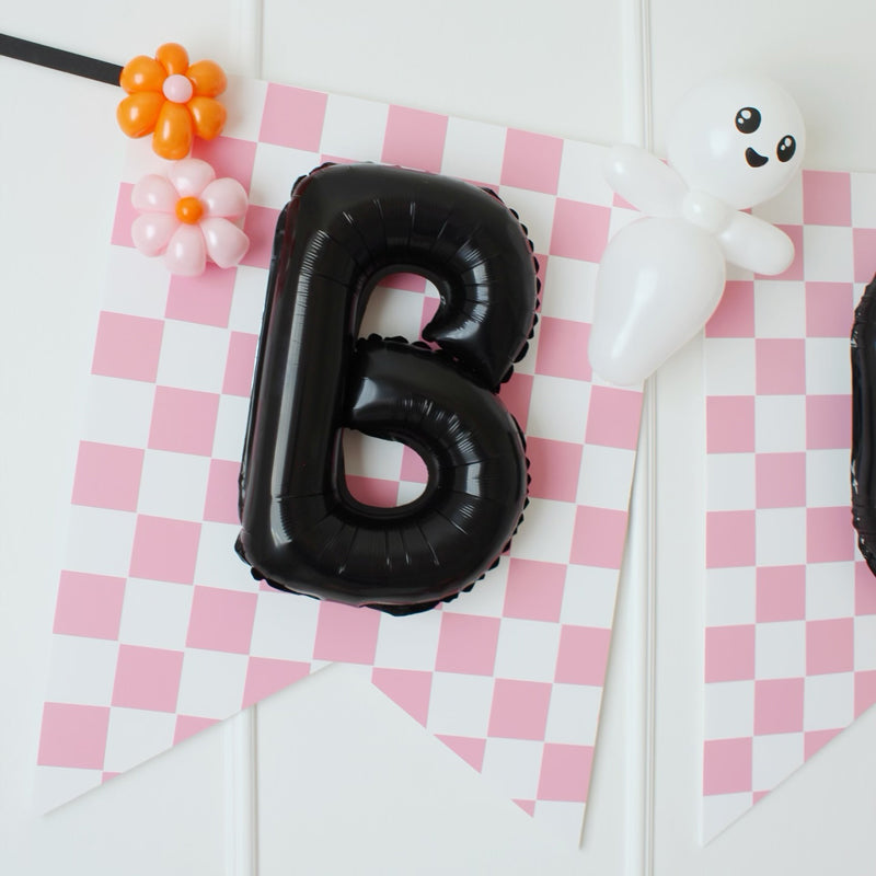A cute mini balloon ghost with a smiling face is attached to a pink and white checkered Halloween banner featuring a black foil letter balloon. Small balloon flowers in orange and pink add a whimsical touch. This festive and playful decoration is perfect for Halloween parties, trick-or-treat setups, and themed events. Part of Lilly Jimenez’s Halloween Balloon Tutorials!