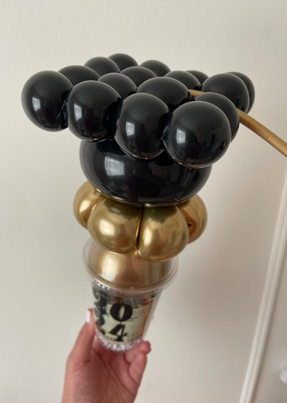 A creative mini graduation cap made from black balloons sits on top of a candy cup filled with gold accents and rolled money. This festive balloon topper is perfect for graduation party favors, gift displays, and photo props. Great for balloon artists, party decorators, and DIY gift makers celebrating the class of 2024.