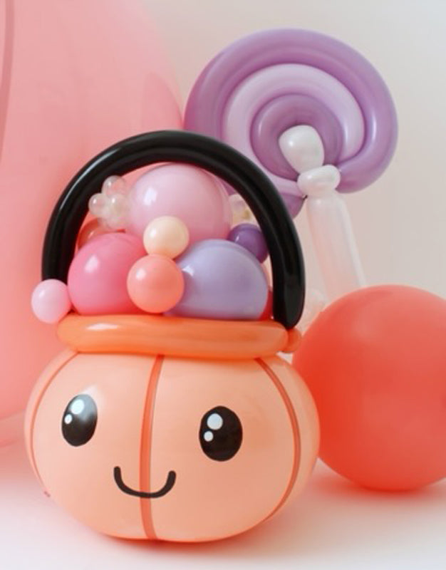 A small balloon pumpkin candy bucket with an adorable smiling face is filled with pastel balloon "candies" and topped with a black balloon handle. A twisted purple balloon lollipop and orange balloon accents complete the festive setup. This whimsical Halloween decoration by Lilly Jimenez of The Creative Heart Studio is perfect for trick-or-treat parties, candy displays, and seasonal decor.