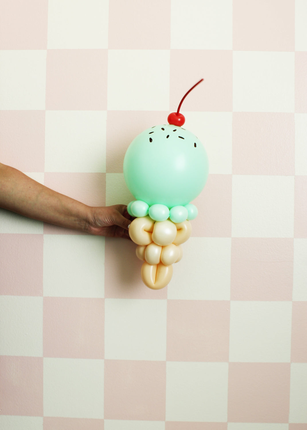Mint green balloon ice cream cone sculpture with a cherry topper and hand-drawn sprinkles, held against a pastel pink checkered wall. Great for summer parties, ice cream-themed events, and balloon decor displays.