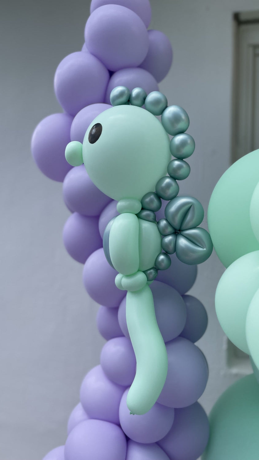 A mint green balloon seahorse sculpture with metallic accents, featured in the Under the Sea Balloon Class by Lilly Jimenez. The seahorse is attached to a pastel purple balloon garland, ideal for ocean, mermaid, or under-the-sea themed parties. This tutorial teaches decorators how to build the seahorse step by step using twisting and layering balloon techniques.