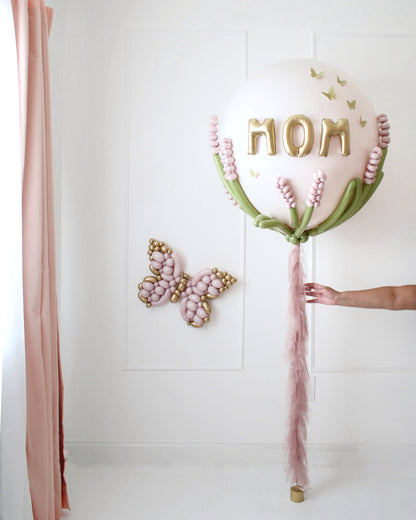 Stunning jumbo Mother's Day balloon with gold "MOM" letters, pastel balloon florals, gold butterflies, and a flowing blush pink tassel. Perfect for Mother’s Day party decor, balloon deliveries, or photo setups.