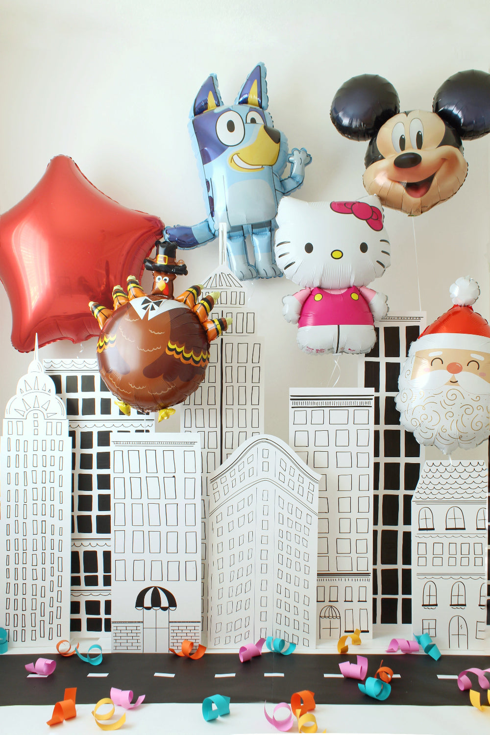 NYC sketch-style building backdrop displayed as a parade-themed party setup with oversized character balloons like Bluey, Mickey, Hello Kitty, a turkey balloon, and a red star floating above the black-and-white city panels. Colorful paper curls decorate the street scene, creating a fun New York parade look perfect for Thanksgiving Day Parade parties, holiday celebrations, and photo backdrops.