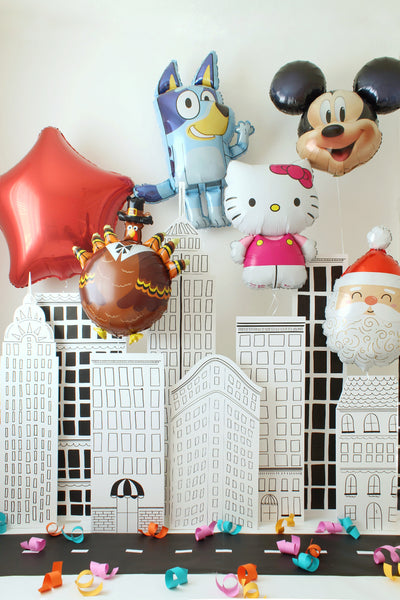 NYC sketch-style building backdrop displayed as a parade-themed party setup with oversized character balloons like Bluey, Mickey, Hello Kitty, a turkey balloon, and a red star floating above the black-and-white city panels. Colorful paper curls decorate the street scene, creating a fun New York parade look perfect for Thanksgiving Day Parade parties, holiday celebrations, and photo backdrops.