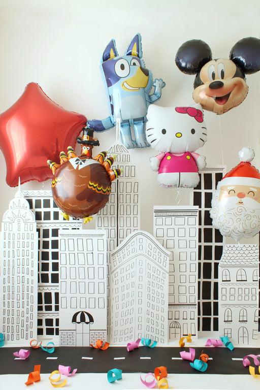 NYC sketch-style building backdrop displayed as a parade-themed party setup with oversized character balloons like Bluey, Mickey, Hello Kitty, a turkey balloon, and a red star floating above the black-and-white city panels. Colorful paper curls decorate the street scene, creating a fun New York parade look perfect for Thanksgiving Day Parade parties, holiday celebrations, and photo backdrops.